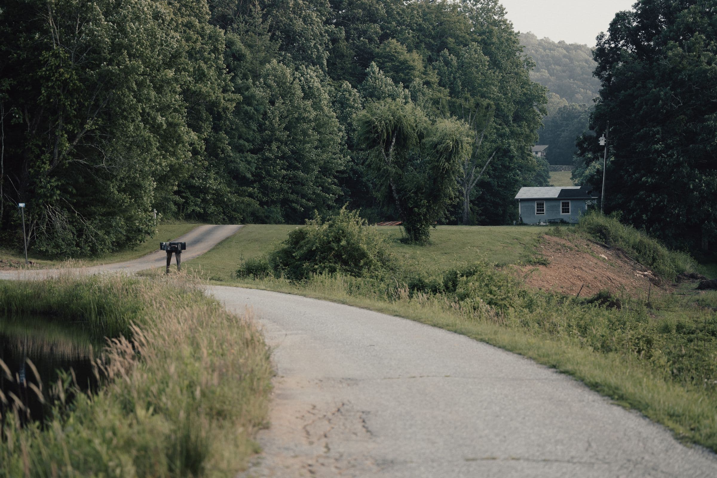 Junk removal in Halls, TN: rural paved road curving past a lawn and a small house with wooded hills behind