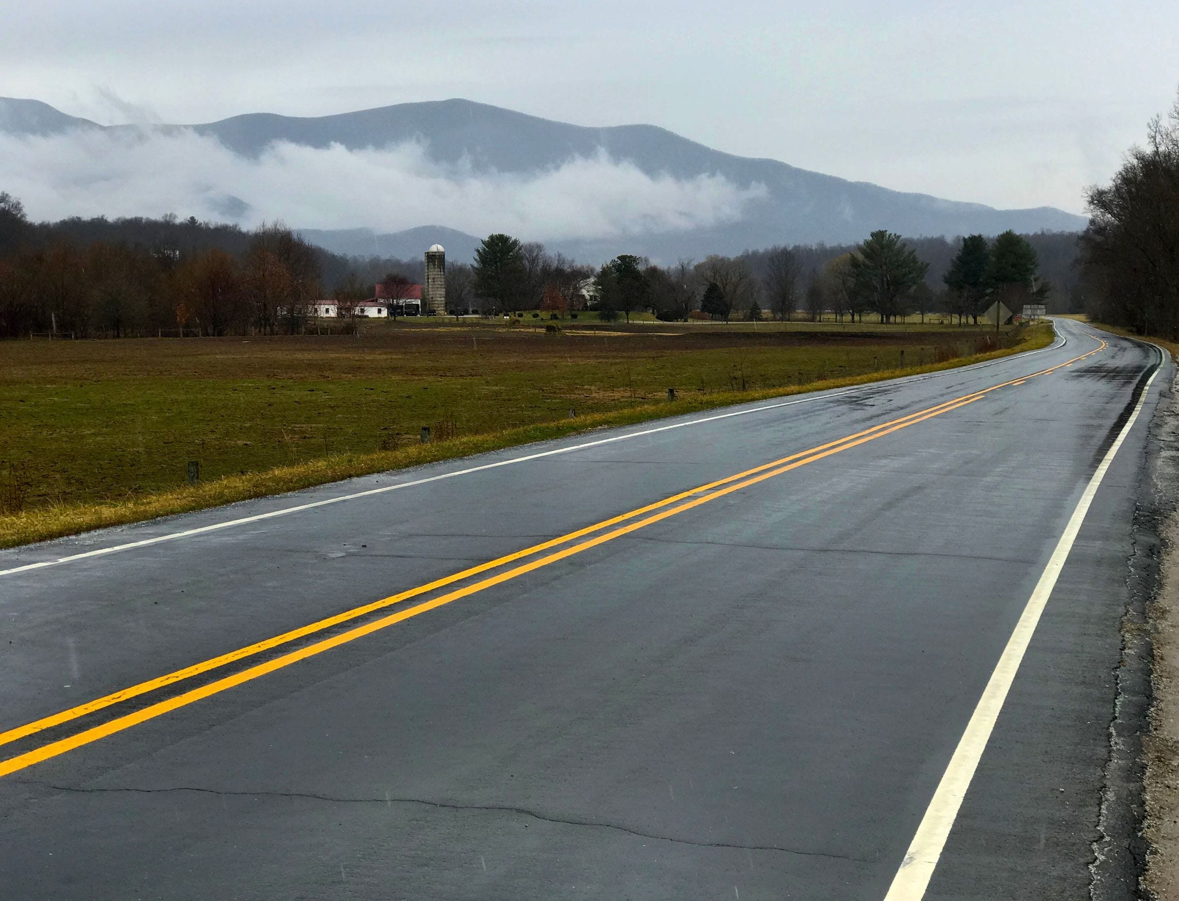 Junk removal in Louisville, TN: two-lane country road past farmland, a silo and farm buildings with low clouds on the mountains